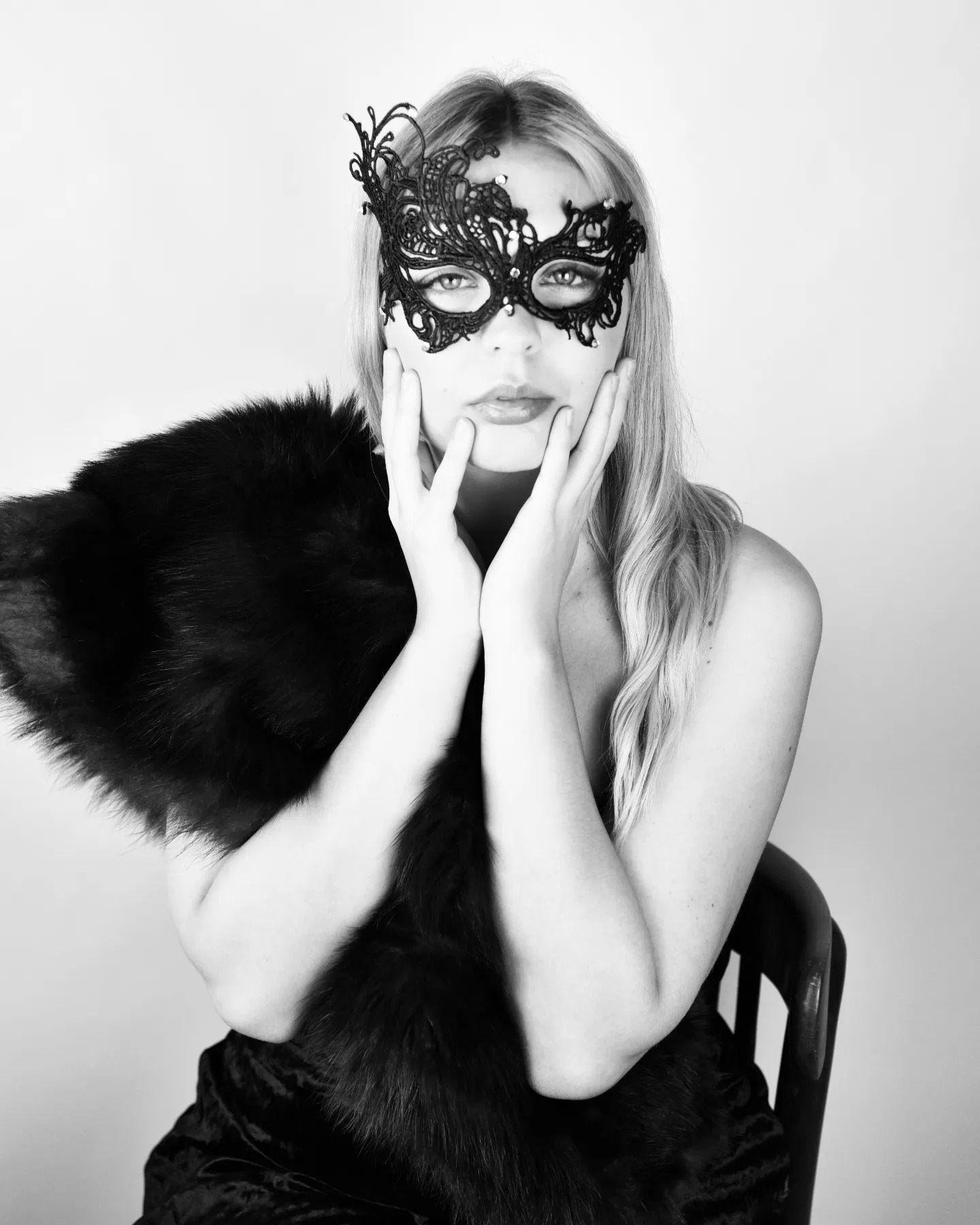 Portret kobiety w czarno-białej tonacji, z koronkową maską na twarzy i futrzanym szalem, opierającej dłonie o policzki, siedzącej na krześle.