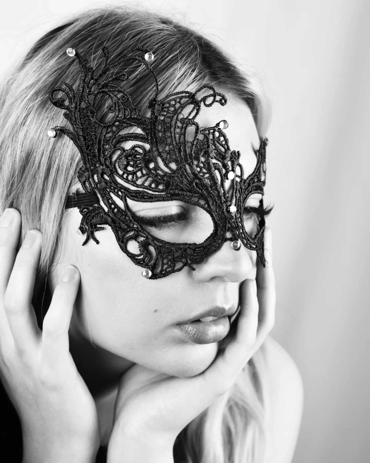 Portret kobiety w czarno-białej tonacji, z koronkową maską na oczach, opierającej brodę na dłoniach, z zamkniętymi oczami i delikatnym makijażem.
