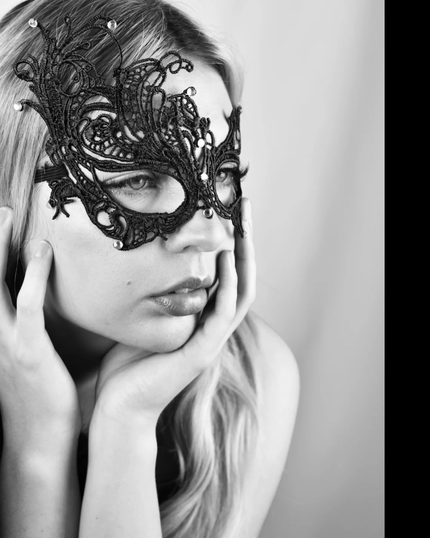 Czarno-białe portretowe ujęcie kobiety w ozdobnej, czarnej, koronkowo-koralikowej masce, opierającej twarz na dłoniach, z zamyślonym spojrzeniem skierowanym w prawo.
