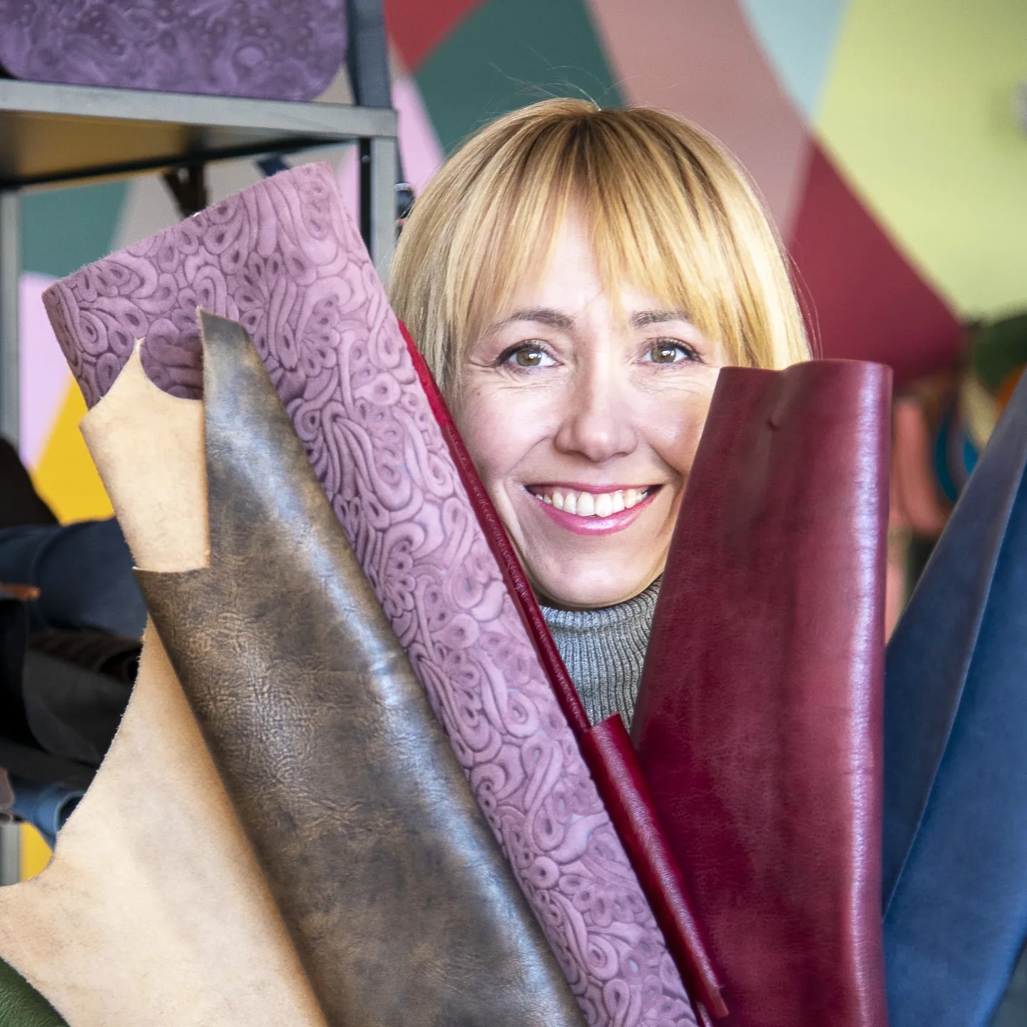 Uśmiechnięta kobieta z blond włosami trzymająca w rękach próbki różnokolorowych skór o różnych fakturach, widoczny regał z wyrobami skórzanymi w tle.