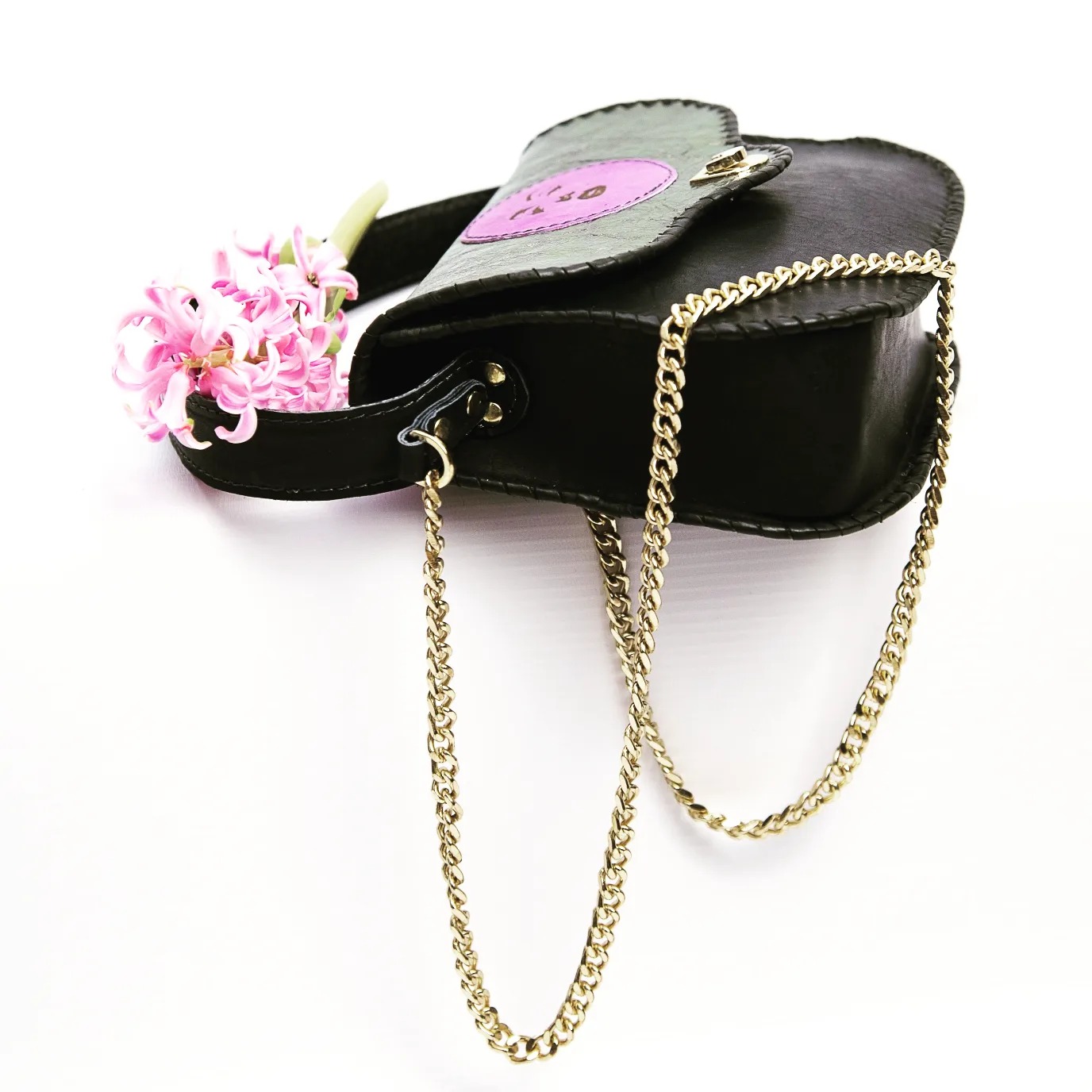 Czarna skórzana torebka z złotym łańcuszkiem i różowym kwiatem hiacyntu w uchwycie na białym tle, widok z góry.