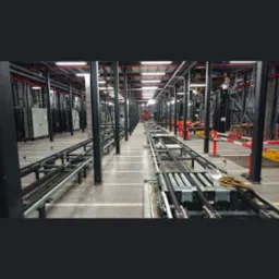 Wnętrze hali produkcyjnej z widoczną linią transportową z rolkami i elementami automatyki, czarne słupy podtrzymujące konstrukcję, czerwone rury pod sufitem, szara betonowa posadzka.