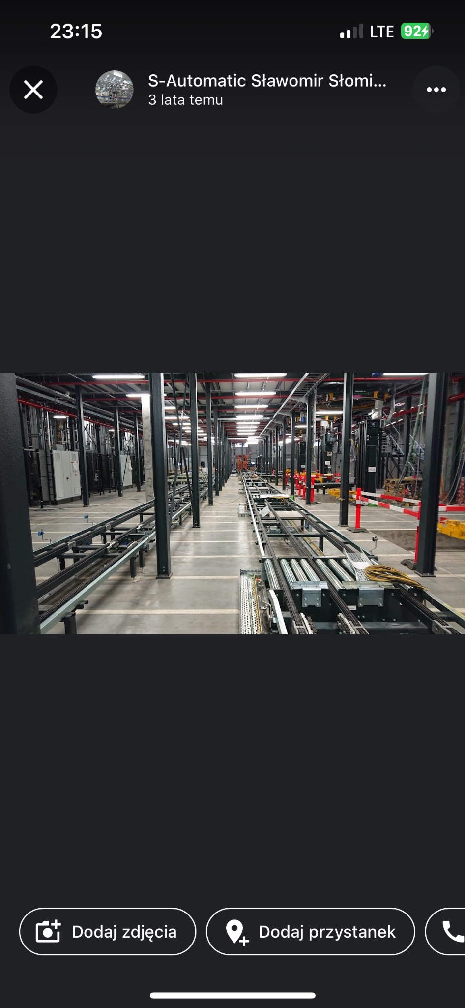 Wnętrze hali produkcyjnej z widoczną linią transportową z rolkami i elementami automatyki, czarne słupy podtrzymujące konstrukcję, czerwone rury pod sufitem, szara betonowa posadzka.