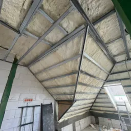 Poddasze w trakcie izolacji, widoczne metalowe profile konstrukcyjne i natryśnięta warstwa izolacyjna o porowatej strukturze, częściowo zakrywająca krokwie dachowe. W tle okno dachowe i fragment...