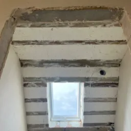 Montaż płyty karton gips na drewnianej konstrukcji sufitu z oknem dachowym, widoczne niedoskonałości i surowe wykończenie.