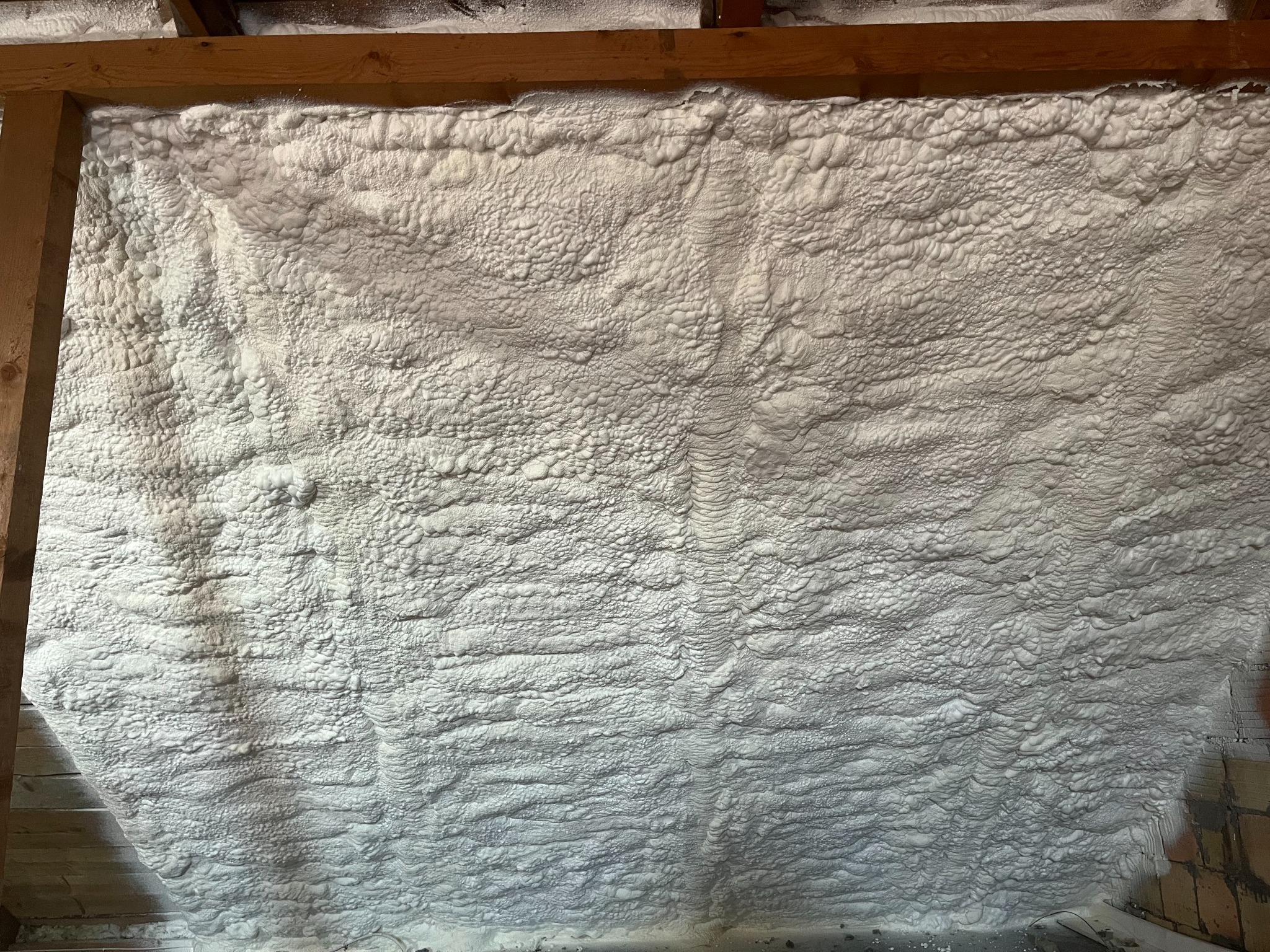 Poddasze ocieplone białą pianką PUR, widoczne drewniane elementy konstrukcyjne dachu i fragment murowanej ściany.