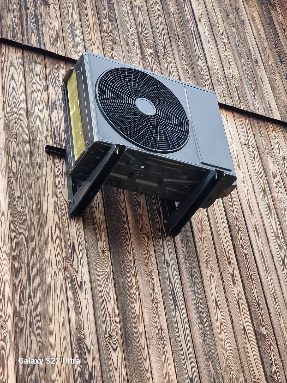 Szara jednostka zewnętrzna klimatyzacji zamontowana na drewnianej elewacji budynku, widoczna z dołu pod kątem.