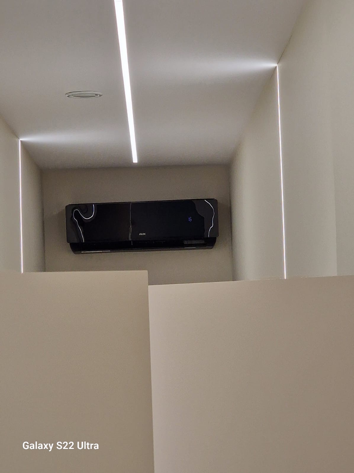 Czarny klimatyzator AUX zamontowany na ścianie, oświetlony linearnymi lampami LED w minimalistycznym wnętrzu z jasnymi ścianami.