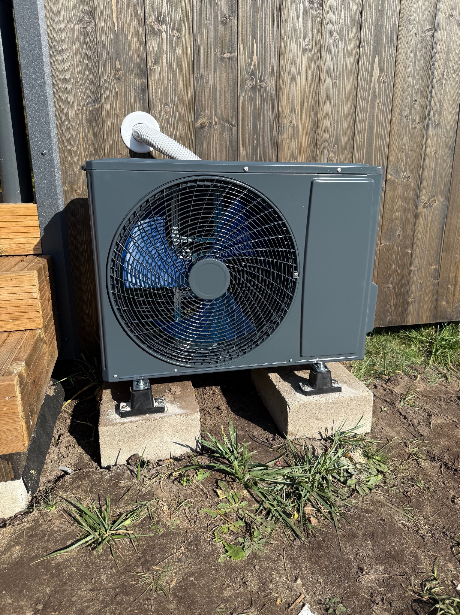 Szara jednostka zewnętrzna klimatyzacji na betonowych bloczkach, rura odprowadzająca skropliny przymocowana do drewnianej ściany, widoczne trawa i ziemia.