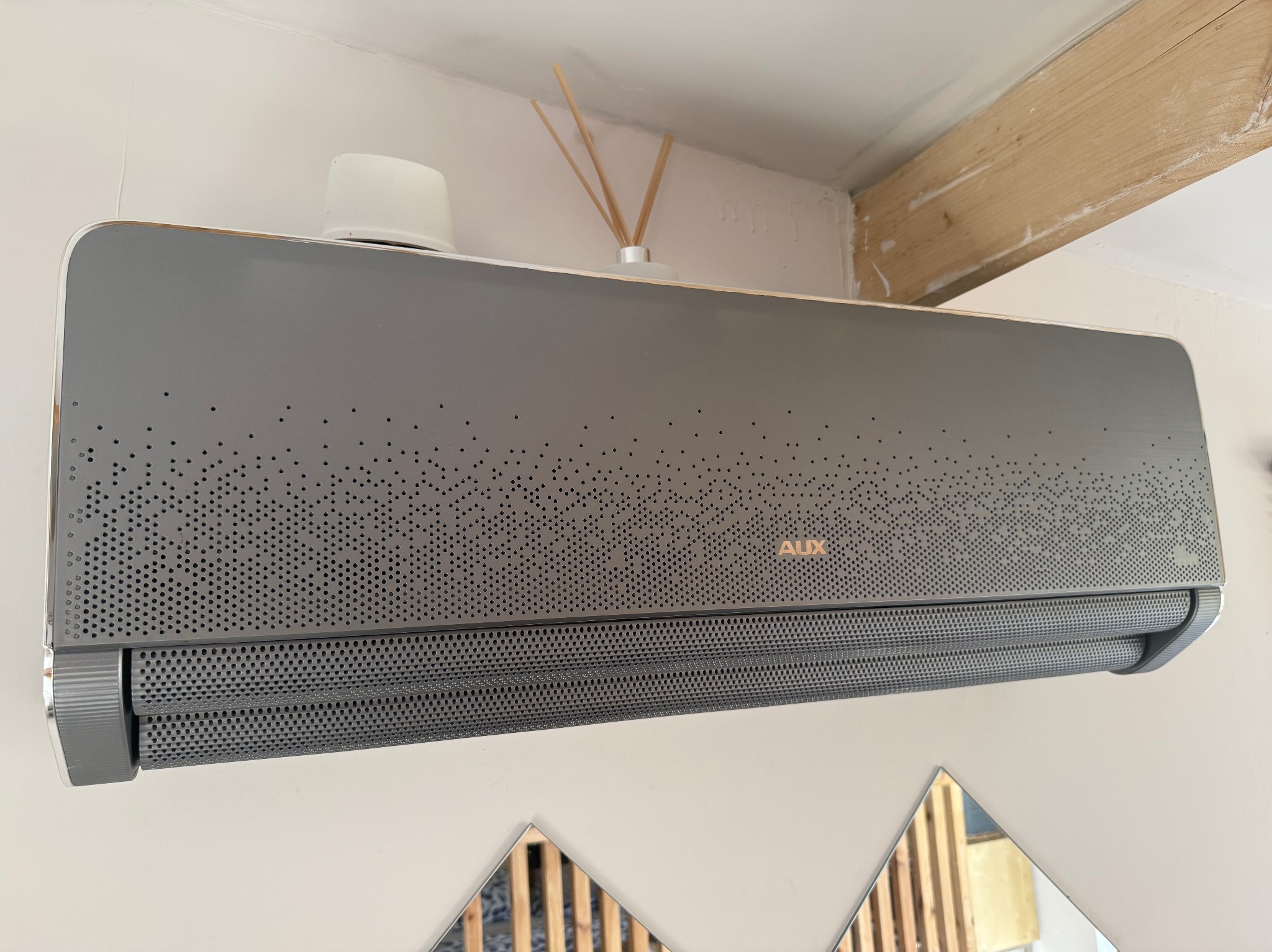 Szary klimatyzator marki AUX zamontowany na białej ścianie, nad nim dekoracyjny dyfuzor zapachowy i drewniana belka stropowa, a pod nim fragment dekoracji ściennej z luster. Widoczne perforacje...