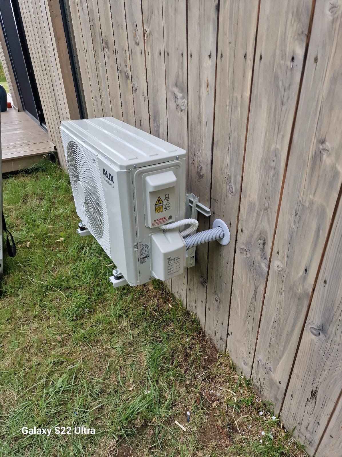 Zewnętrzna jednostka klimatyzacji AUX zamontowana na drewnianej ścianie budynku, widoczne przewody i elementy mocujące.