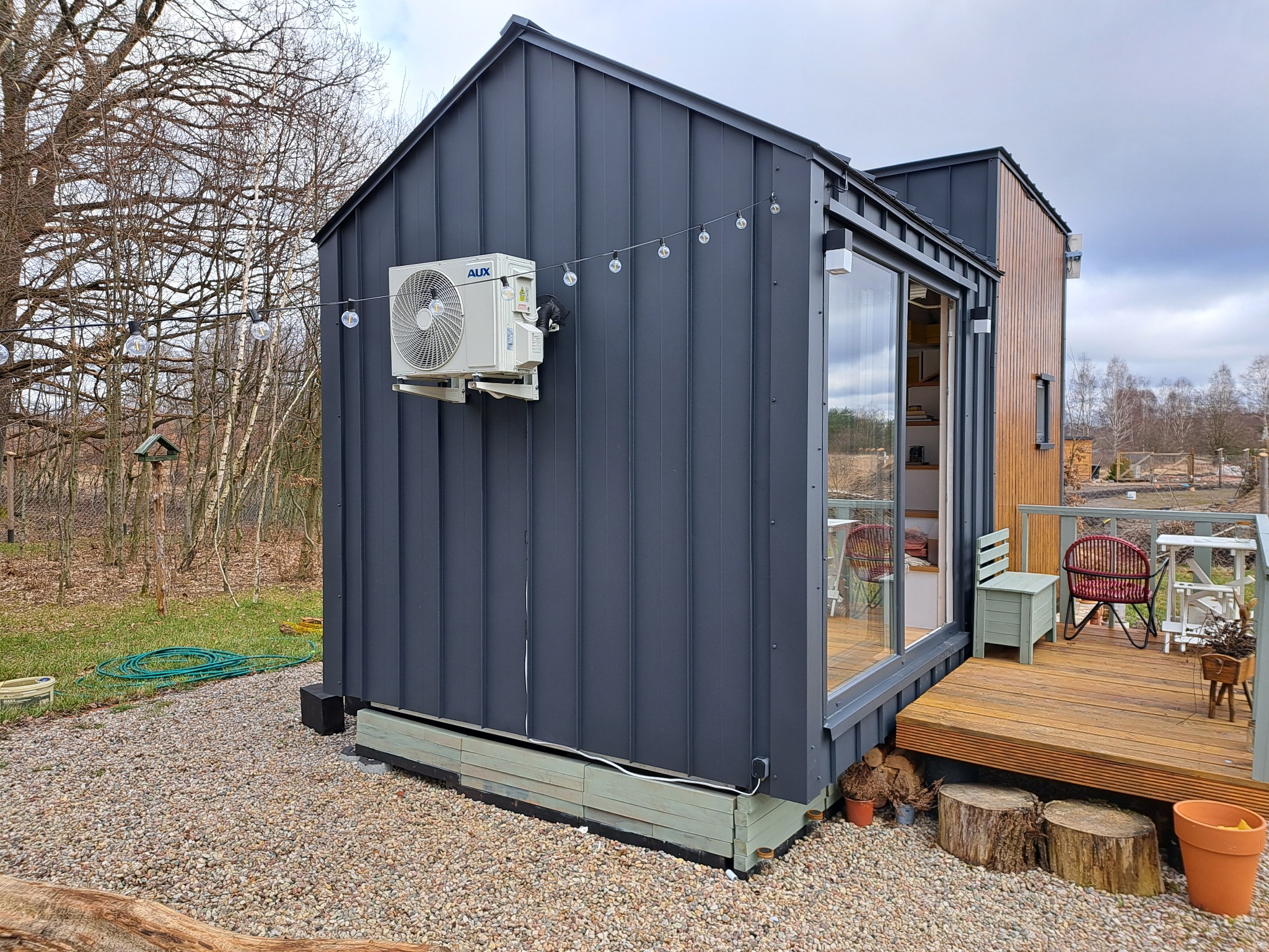 Nowoczesny, ciemnoszary domek z klimatyzacją AUX na elewacji, oświetlony girlandą żarówek, z drewnianym tarasem i widokiem na ogród.