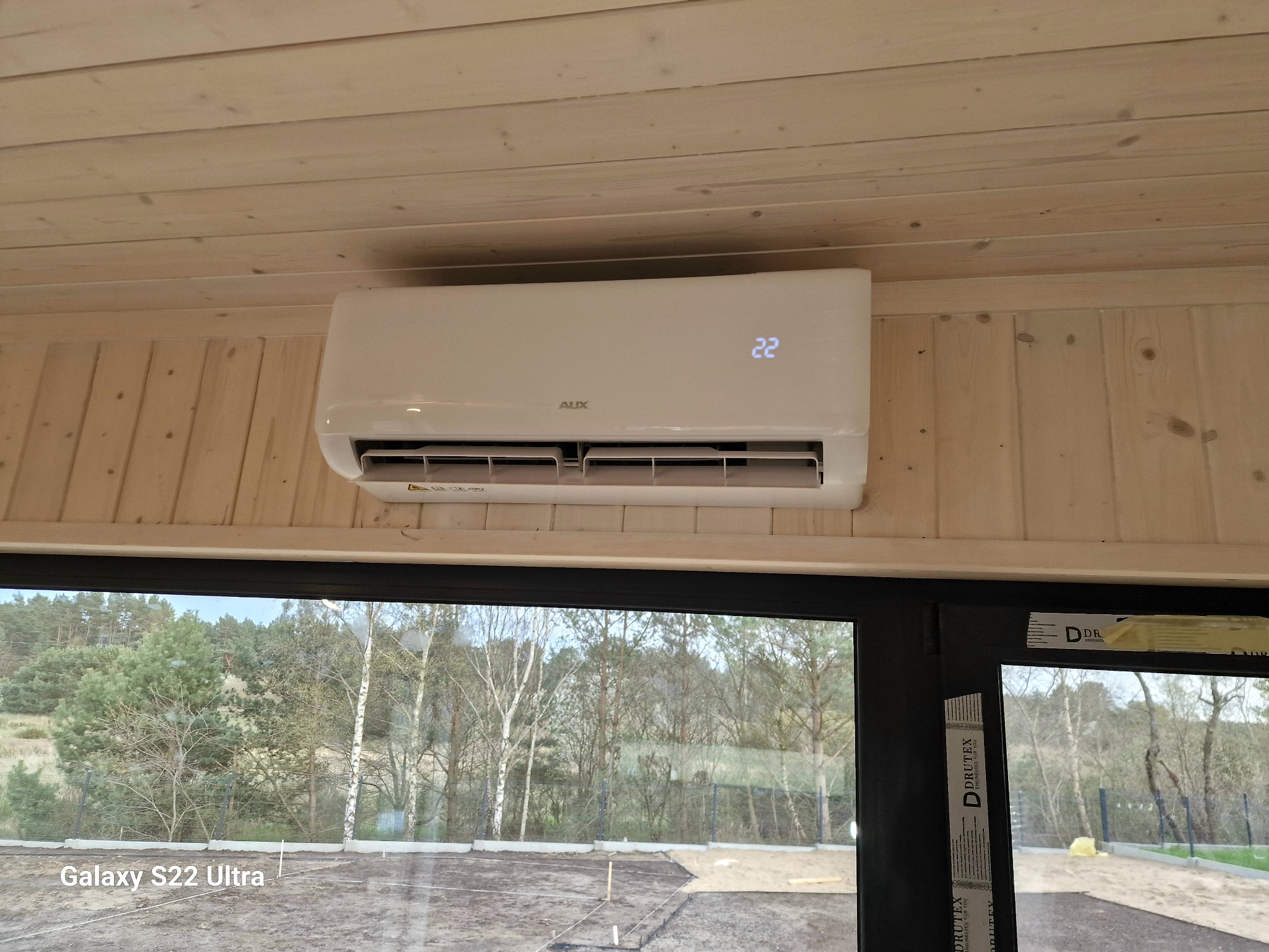 Biały klimatyzator marki AUX zamontowany na drewnianej ścianie w nowym domu, widok przez duże okno na krajobraz w trakcie budowy.