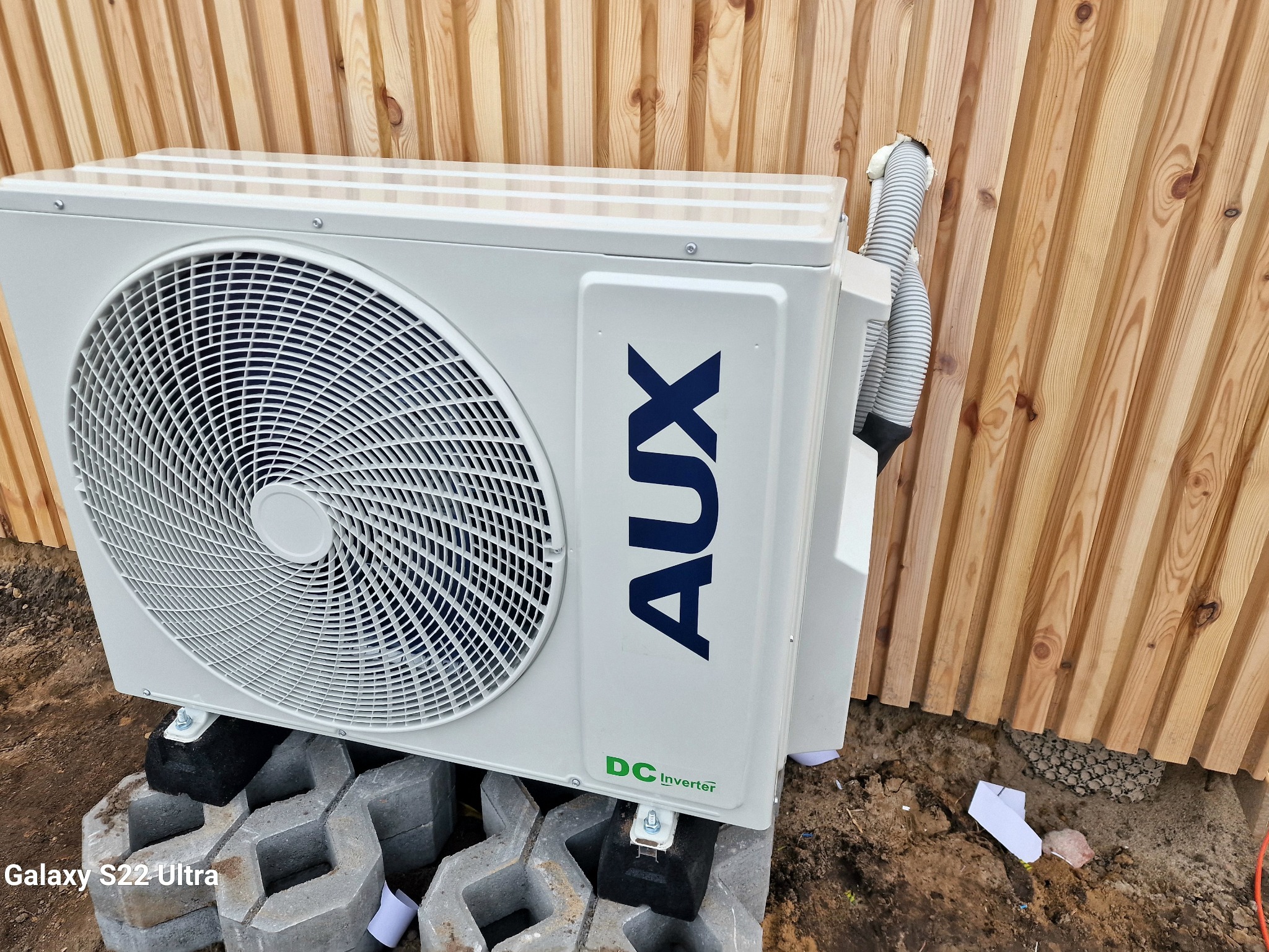 Zewnętrzna jednostka klimatyzacji AUX DC Inverter, zamontowana na betonowych bloczkach, przy drewnianej elewacji z widocznym przewodem wychodzącym przez otwór w desce.