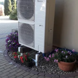 Zewnętrzna jednostka pompy ciepła Panasonic Inverter zamontowana na białym stelażu, otoczona kamieniami ozdobnymi i kwitnącymi kwiatami w donicach, na tle elewacji domu i brukowanego chodnika.