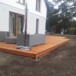 Nowo wybudowany drewniany taras przy domu z szarymi meblami ogrodowymi i rozłożonym parasolem w tle, widoczny fragment ogrodu i osoba.