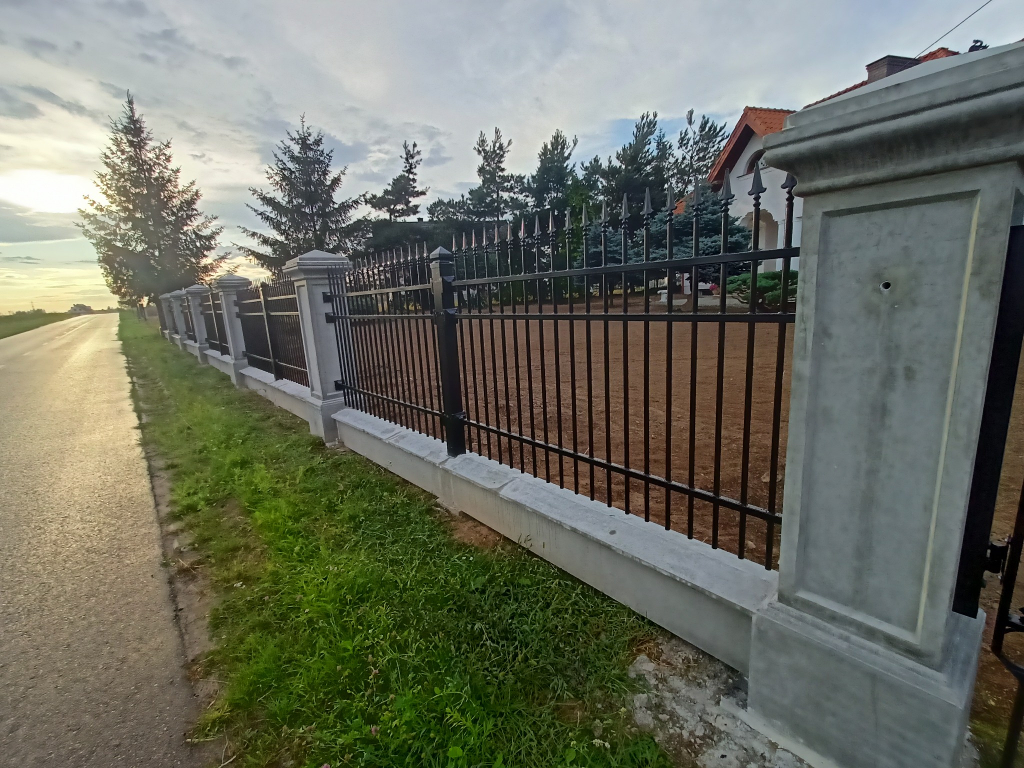 Metalowe ogrodzenie z betonowymi słupkami i podmurówką, widok z boku, trawa i droga wzdłuż ogrodzenia, dom w tle, pochmurne niebo o zachodzie słońca.
