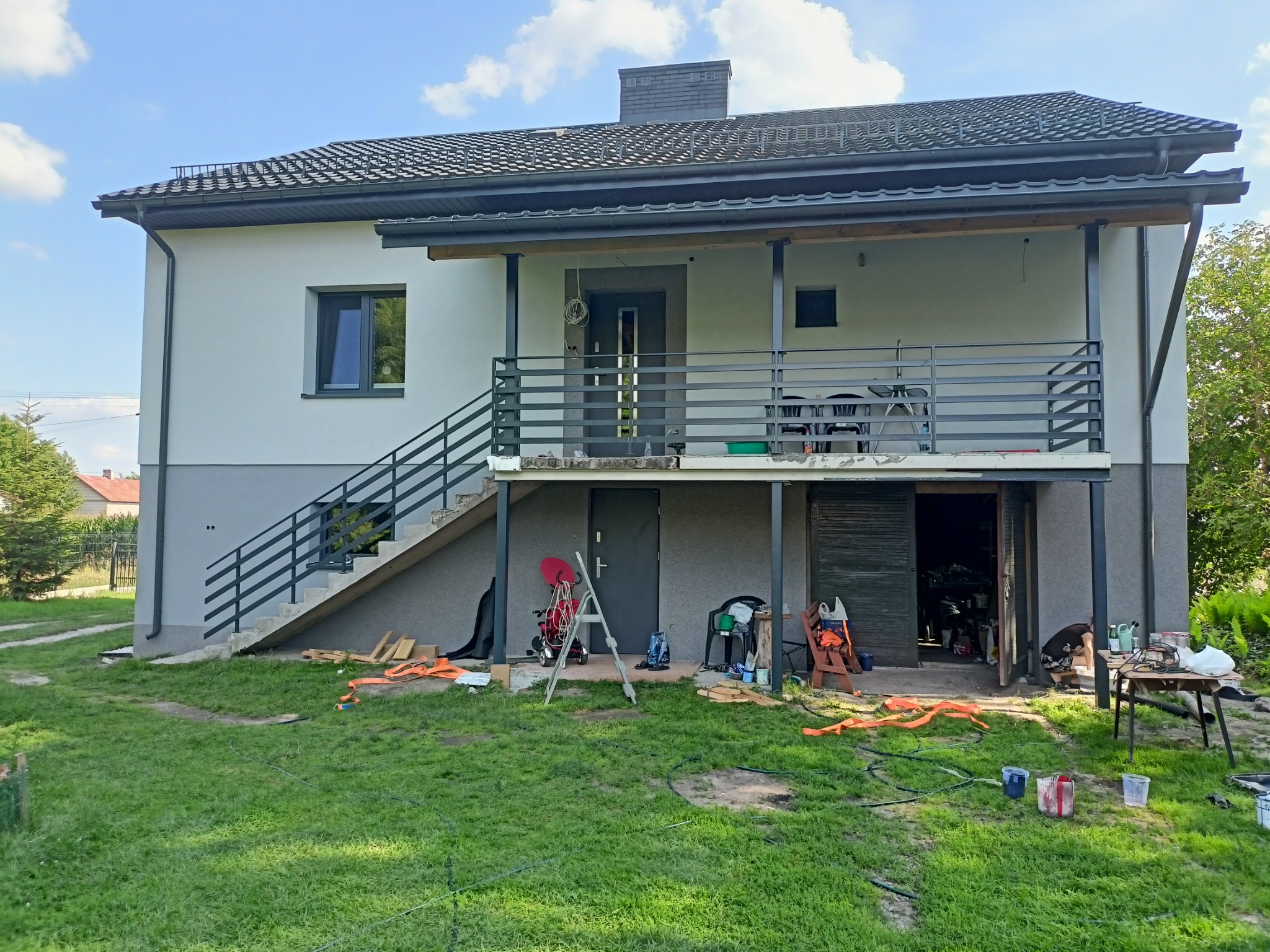 Dwukondygnacyjny dom z szarymi metalowymi schodami zewnętrznymi i balustradą. Na trawniku narzędzia i farby, sugerujące prace wykończeniowe.