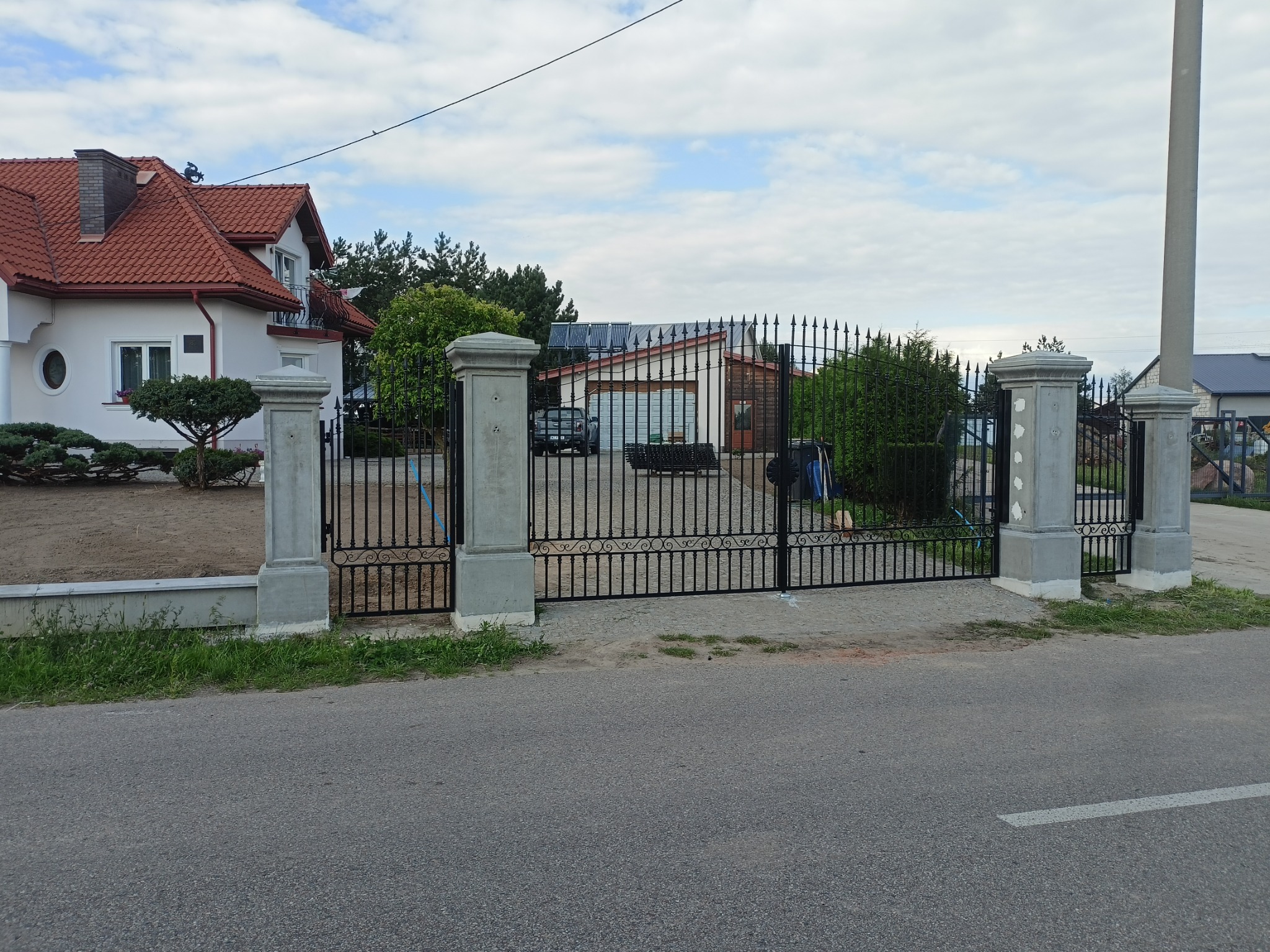 Czarna, metalowa brama wjazdowa z ozdobnymi elementami, osadzona na betonowych słupkach, na tle domu z czerwoną dachówką i zieleni. Widok z ulicy.