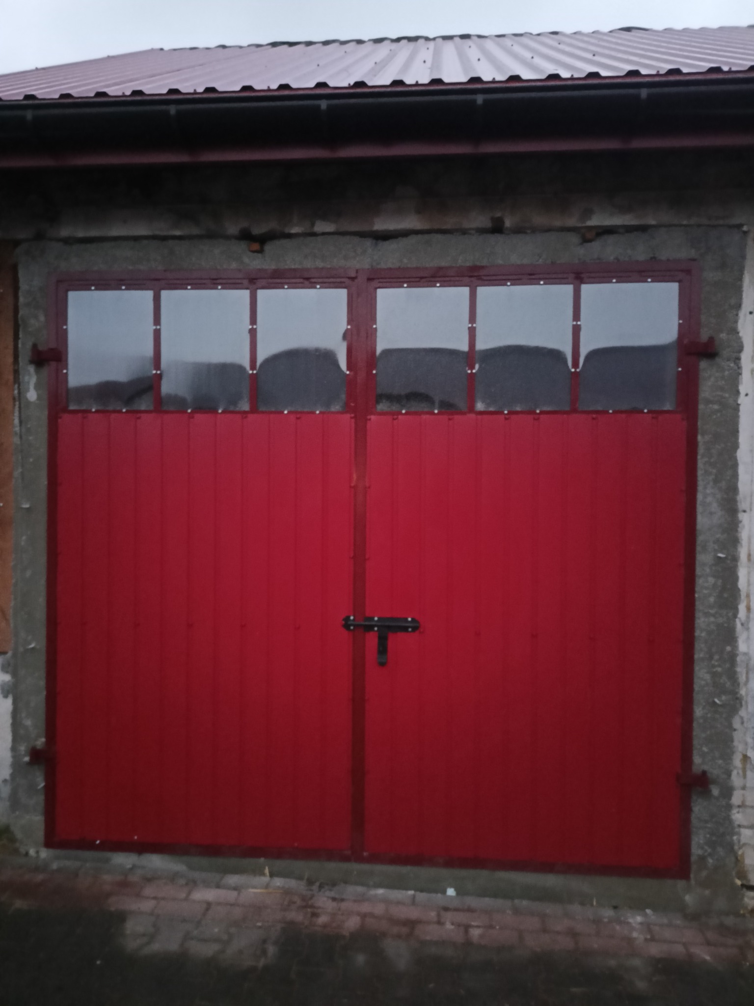 Czerwona, dwuskrzydłowa brama garażowa z przeszklonymi panelami górnymi, zamontowana w szarej, betonowej ścianie budynku. Widoczne ślady wilgoci na dole.