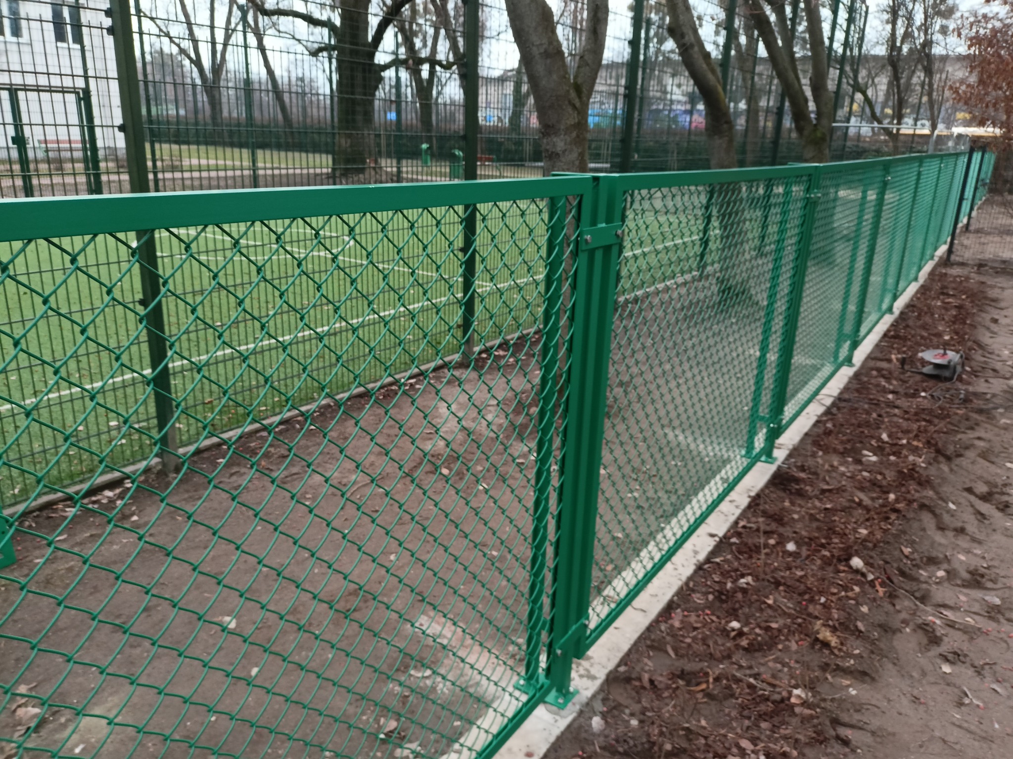 Nowe, zielone ogrodzenie z siatki, otaczające boisko sportowe. Widoczne słupki i siatka, ziemia wokół. Ujęcie z boku, z bliska, prezentujące solidną konstrukcję.