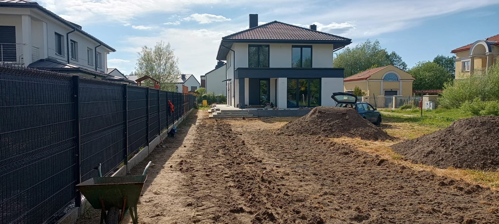 Nowoczesny dom z czarnym ogrodzeniem panelowym. Przygotowany teren pod trawnik, widoczna taczka i pryzmy ziemi. Słoneczny dzień.