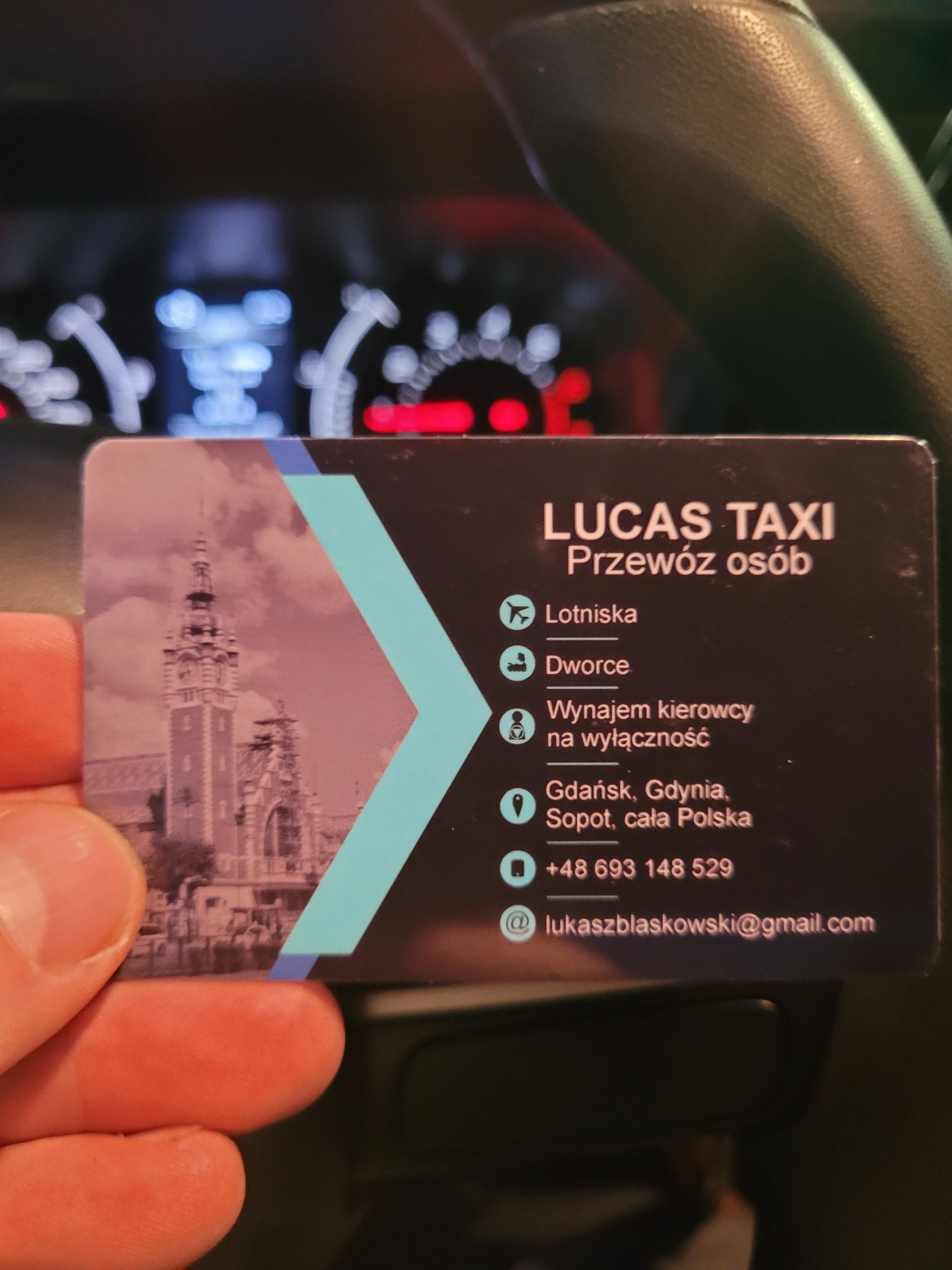 Wnętrze samochodu z widoczną kartą wizytową Lucas Taxi oferującą przewozy osobowe, wynajem kierowcy, z adresem e-mail i numerem telefonu.