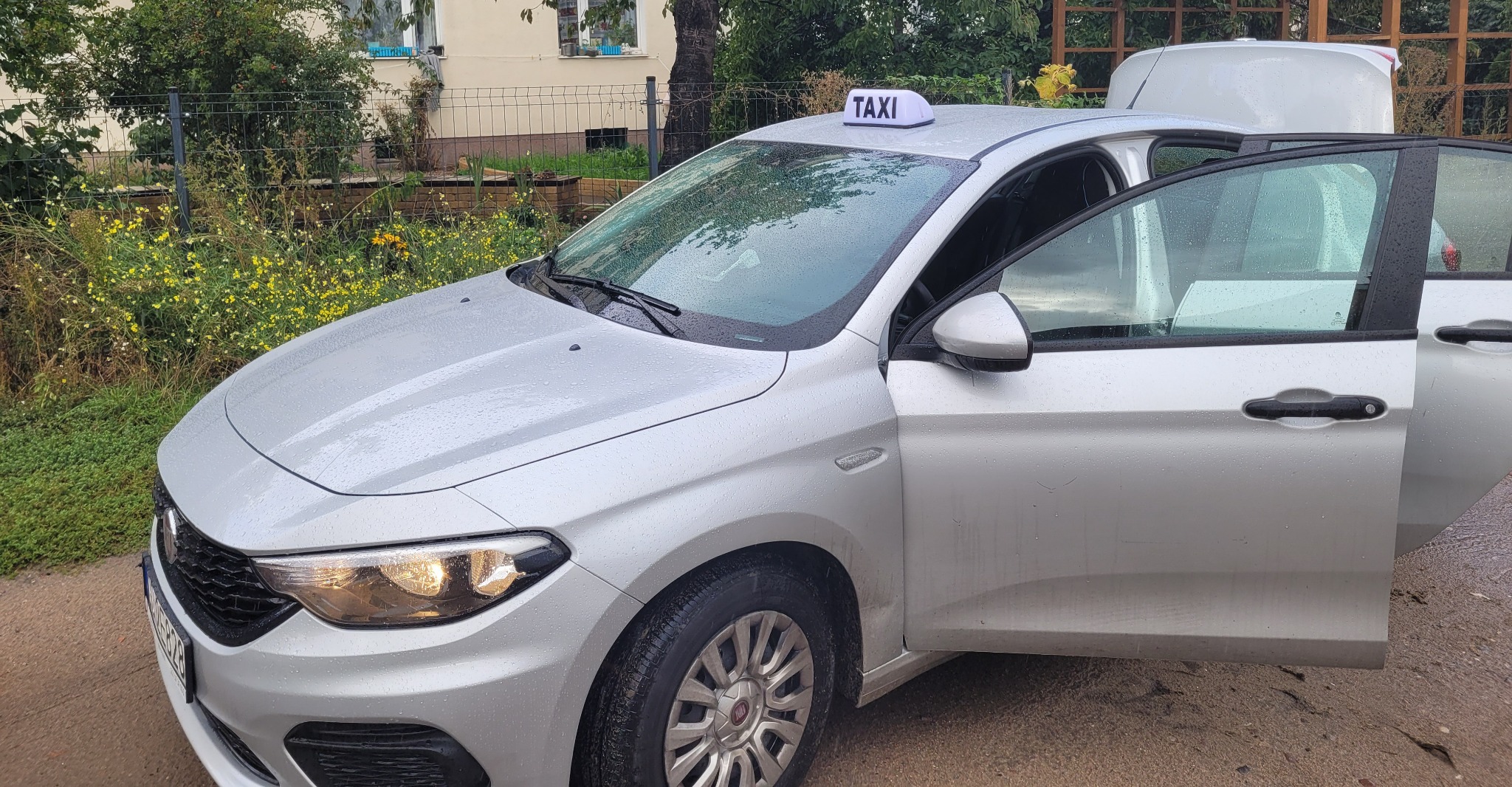 Srebrny samochód taxi z otwartymi drzwiami na zewnątrz, mokry od deszczu, z podświetlonym napisem TAXI na dachu.