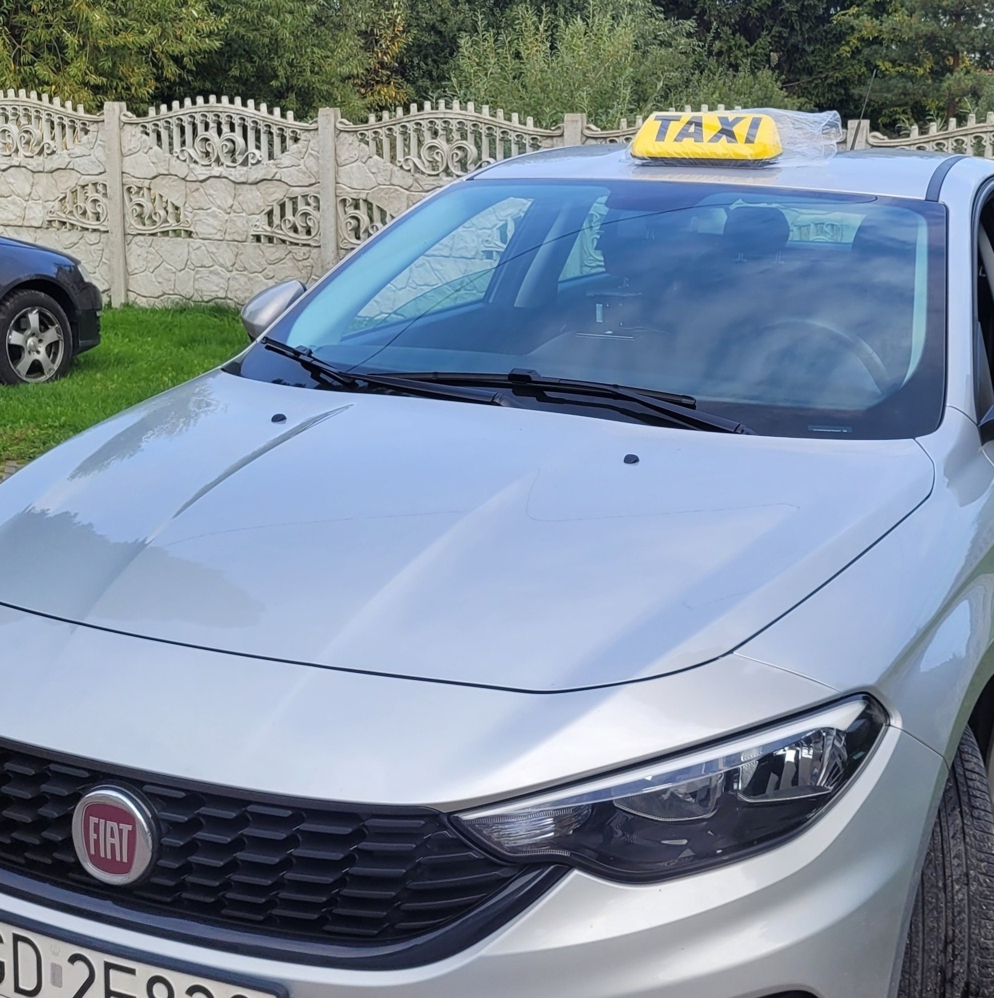 Srebrny Fiat Tipo z żółtym szyldem TAXI na dachu, zaparkowany przed ozdobnym, betonowym ogrodzeniem. Na szyldzie widoczna folia ochronna.