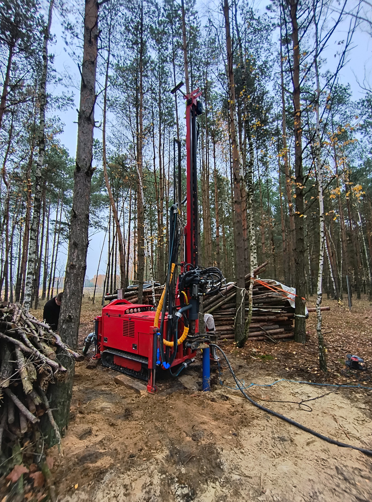 Czerwona wiertnica gąsienicowa w lesie podczas wiercenia studni głębinowej w Skrzydłowicach. Widoczne drzewa i przygotowane drewno.