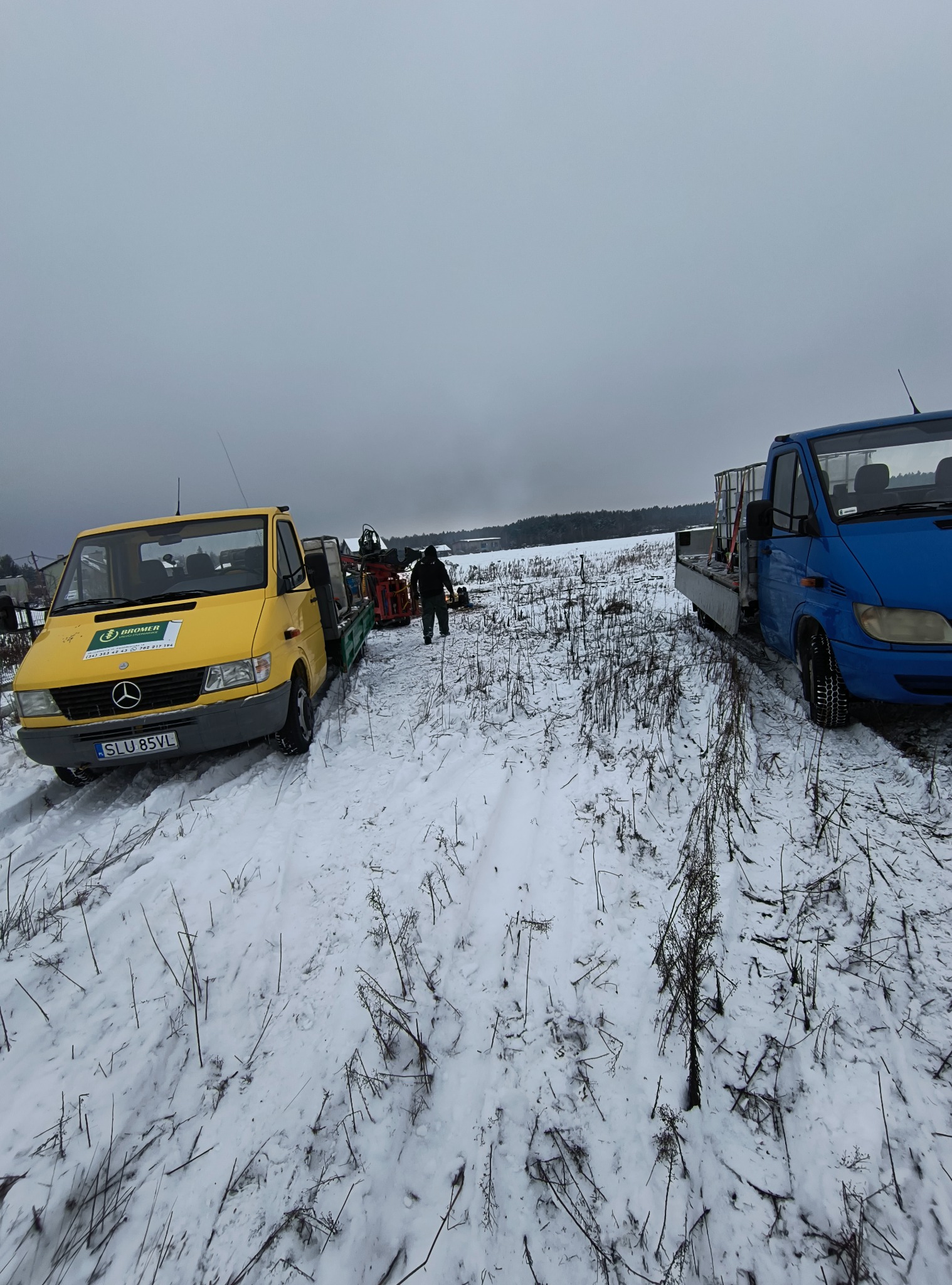 Dwa samochody ciężarowe (żółty i niebieski) na zaśnieżonym terenie, z maszyną wiertniczą na lawecie i osobą w oddali. Pochmurne niebo.