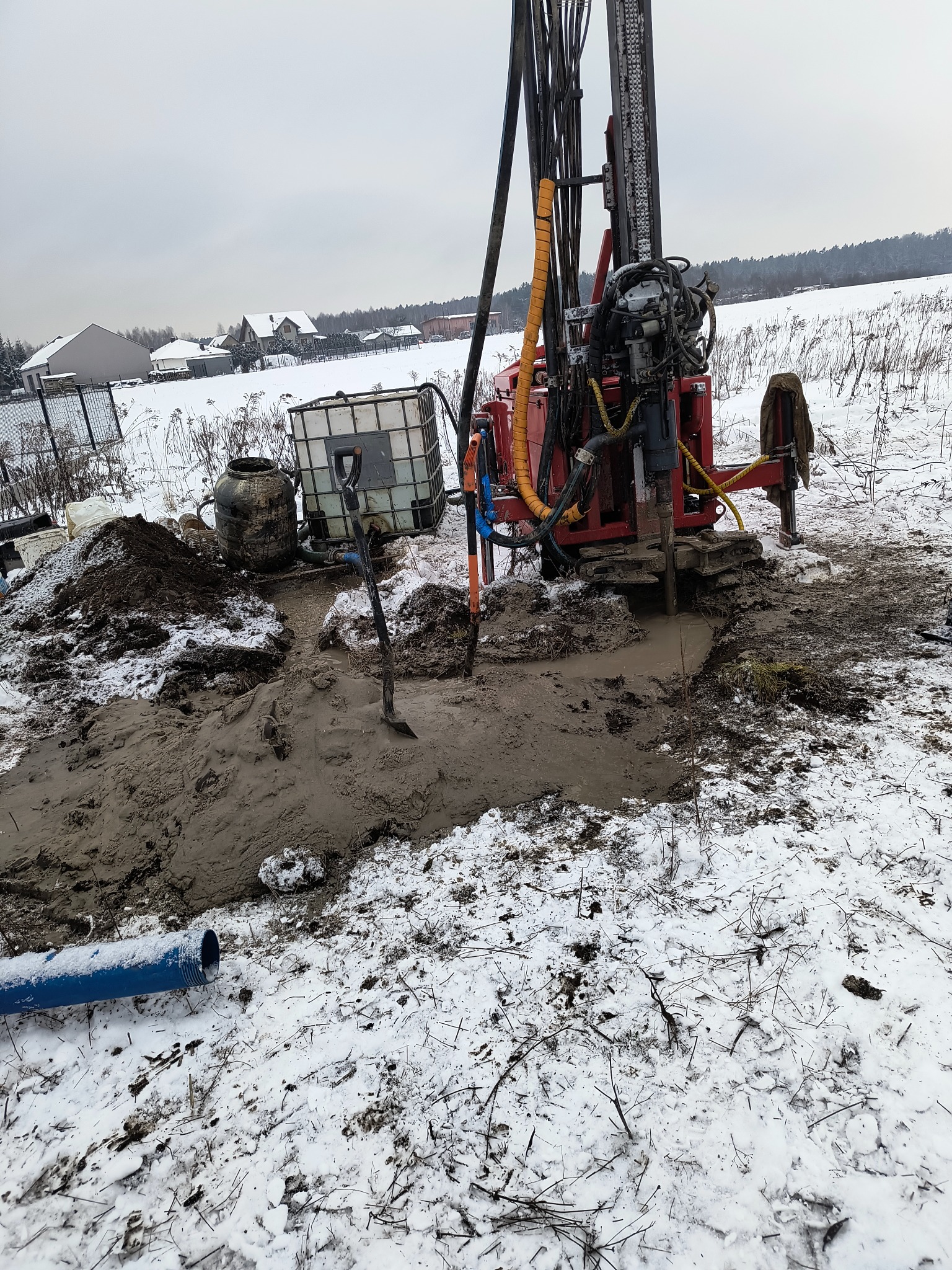 Wiercenie studni głębinowej w zimowym krajobrazie. Czerwona wiertnica na gąsienicach, obok zbiornik na wodę i wykop. Śnieg pokrywa ziemię, w tle zabudowania.