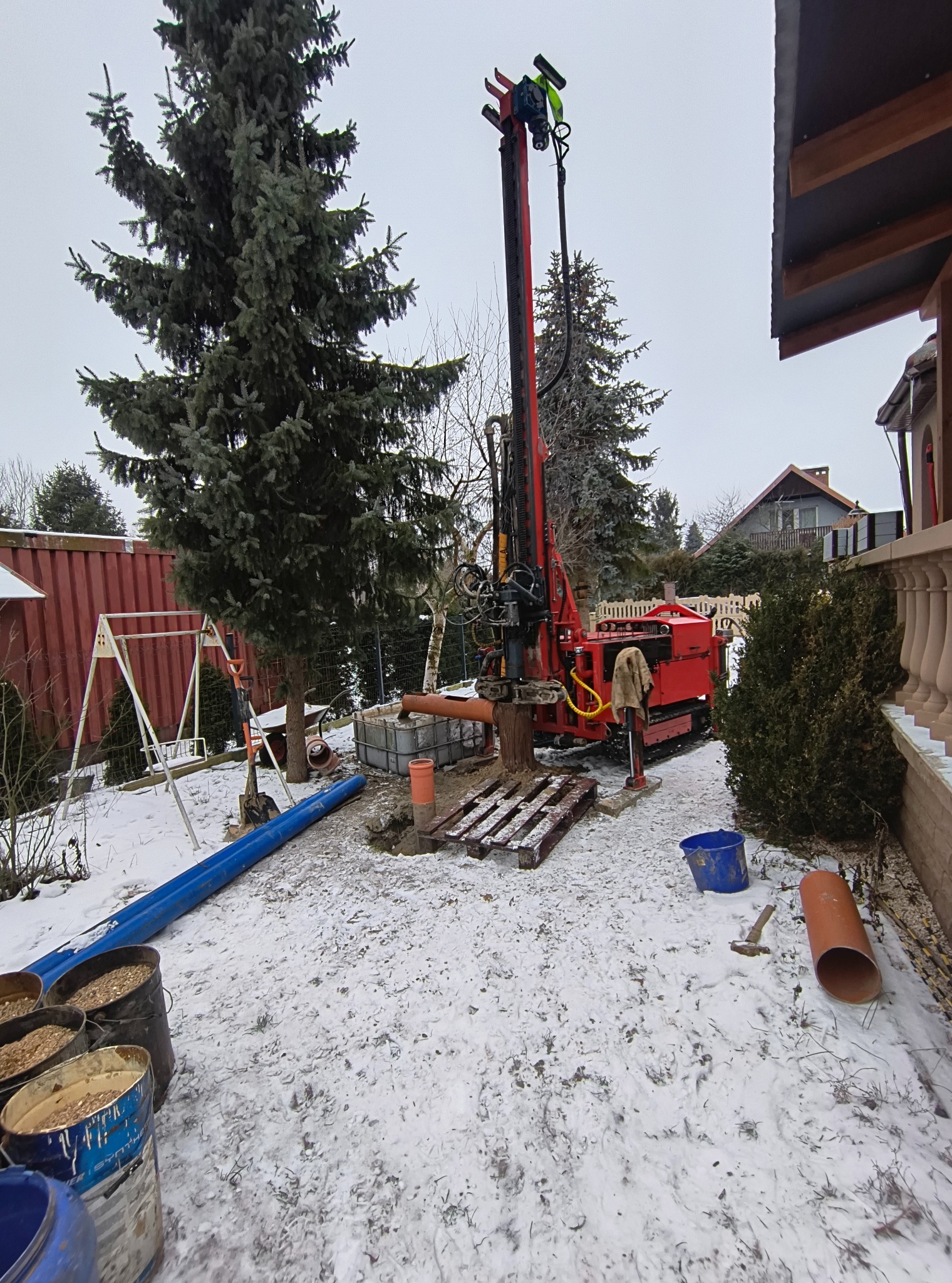 Czerwona wiertnica na śniegu, przygotowana do wiercenia studni. Widoczne rury, palety i pojemniki. W tle dom i drzewa iglaste.
