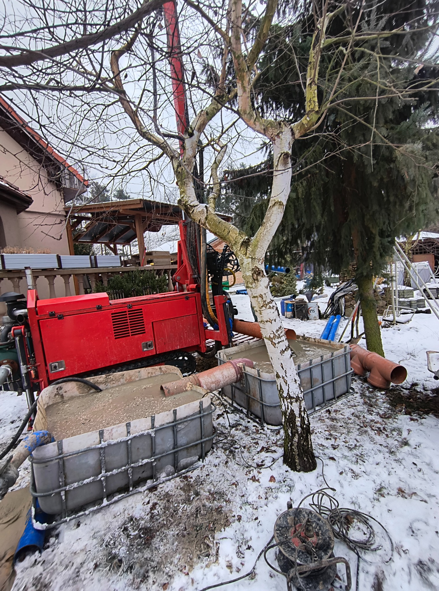 Czerwona wiertnica na śniegu, obok drzewo i zbiorniki z szarą cieczą. W tle dom z drewnianą altaną. Zimowa sceneria, sprzęt do wiercenia studni.