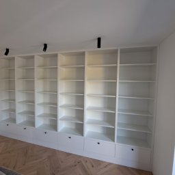 Stolator - Biała, zabudowana szafa z półkami i szufladami, oświetlona punktowymi lampami sufitowymi, na tle parkietu ułożonego w jodełkę.