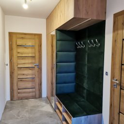 Stolator - Przedpokój z zabudową meblową w odcieniach drewna i zieleni, z miękkim siedziskiem i wieszakami, oświetlony pojedynczą żarówką na suficie i wykończony płytkami imitującymi marmur.