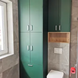 Wysoka, zielona szafa łazienkowa z szufladą i srebrnymi uchwytami, zamontowana obok toalety i drewnianej półki w nowoczesnej łazience wykończonej płytkami imitującymi marmur.