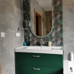 Nowoczesna łazienka z zieloną szafką pod umywalką, owalnym lustrem i geometryczną tapetą w odcieniach szarości, zieleni i brązu.