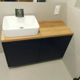 Nowoczesna łazienka z umywalką nablatową, drewnianym blatem i szafką w kolorze granatowym, minimalistyczna armatura, jasne płytki na ścianach i podłodze.