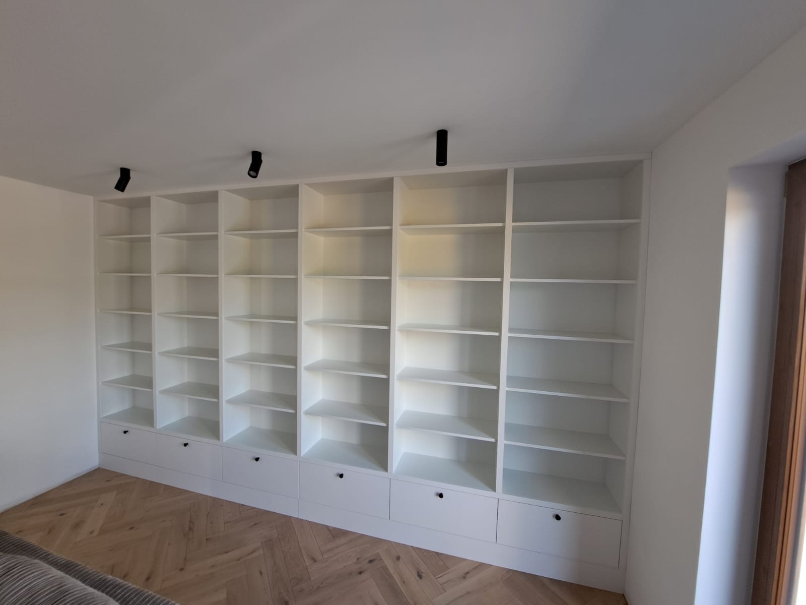 Biała, zabudowana szafa z półkami i szufladami, oświetlona punktowymi lampami sufitowymi, na tle parkietu ułożonego w jodełkę.