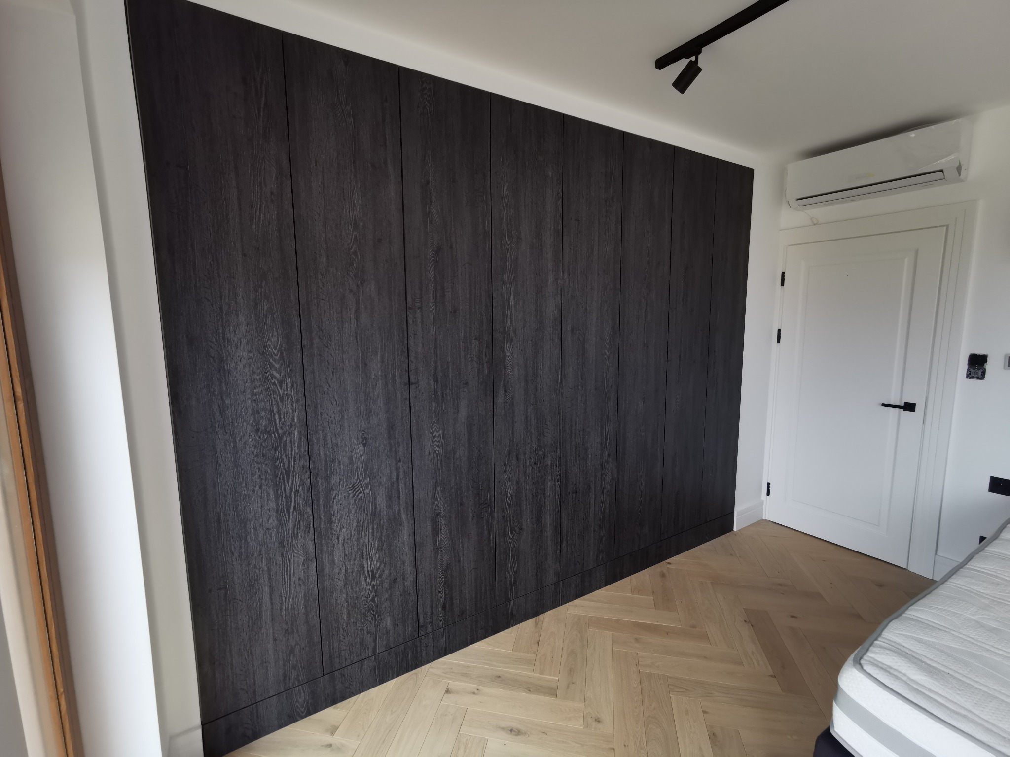 Duża, ciemna szafa wnękowa z drewnianymi frontami, parkiet ułożony w jodełkę, białe ściany i drzwi w tle.