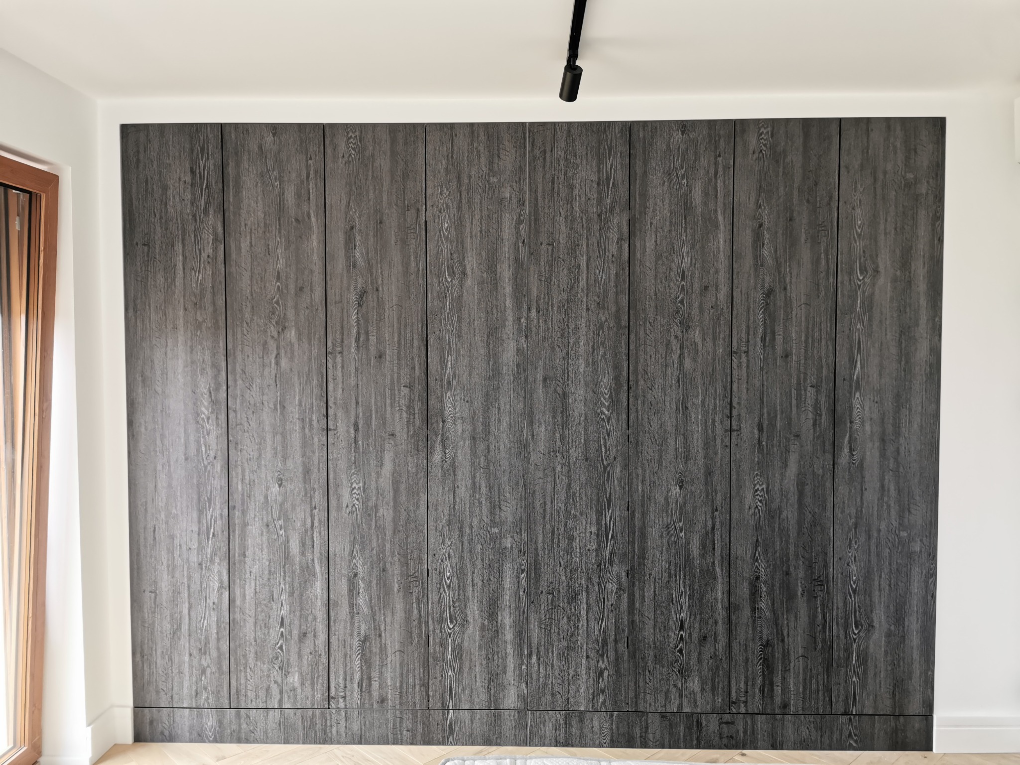 Duża, zabudowana szafa z ciemnego drewna z widocznym usłojeniem, zajmująca całą ścianę, z subtelnymi, ukrytymi uchwytami, oświetlona punktowym reflektorem sufitowym.