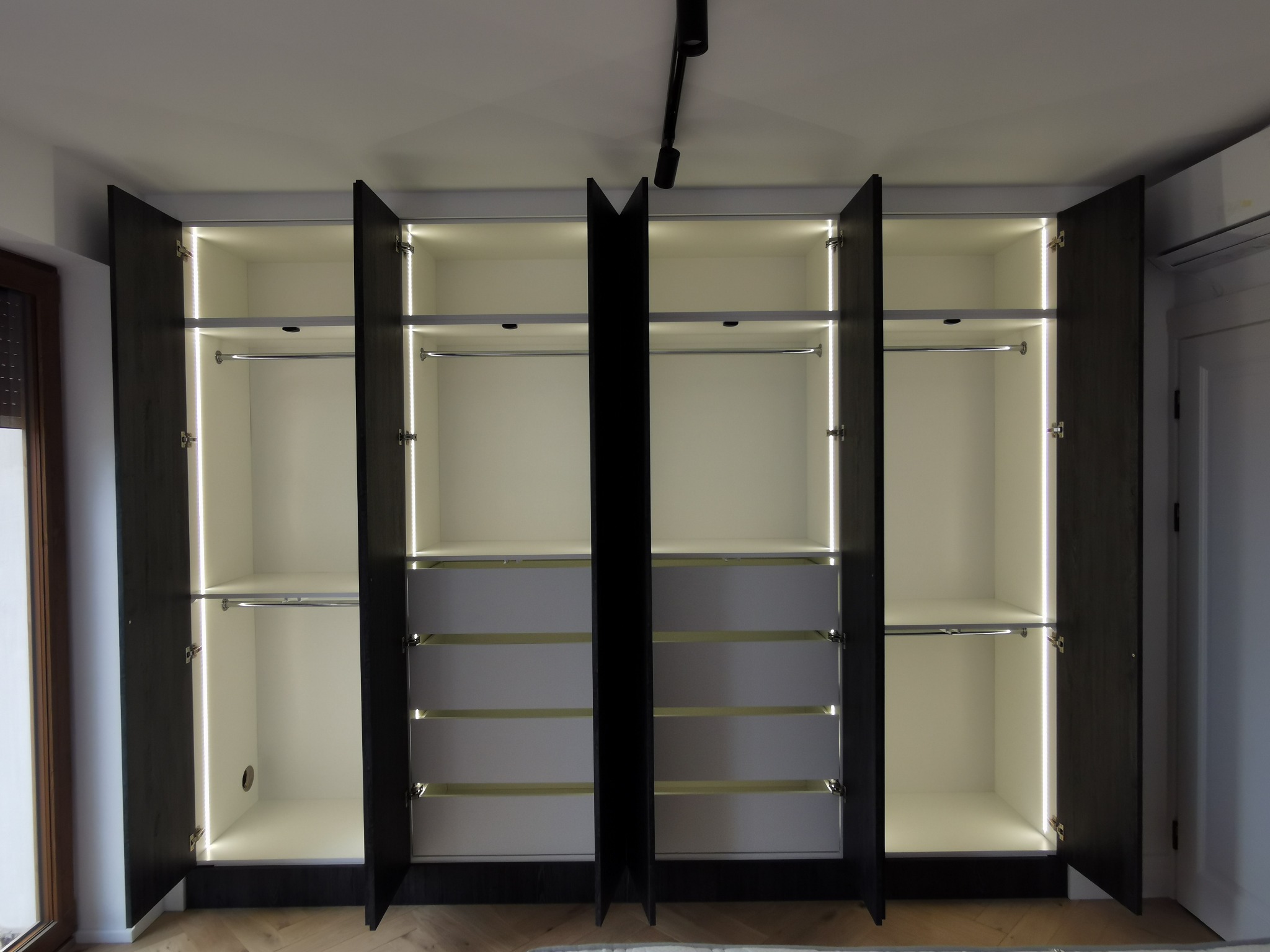 Otwarta, duża szafa z ciemnymi drzwiami i białym wnętrzem, oświetlona pionowymi paskami LED, z drążkami na ubrania i szufladami, widok na drewnianą podłogę w jodełkę.