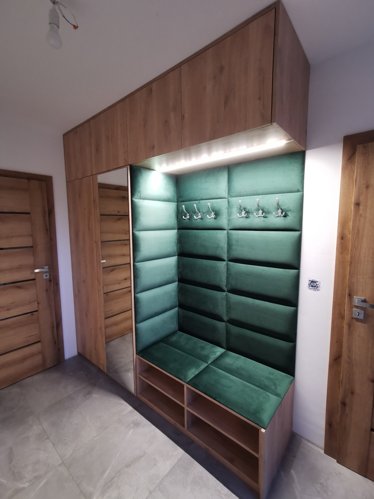 Wnętrze przedpokoju z zabudową meblową z drewnianymi szafkami górnymi, lustrem i tapicerowanym siedziskiem w kolorze butelkowej zieleni z wieszakami na ubrania.