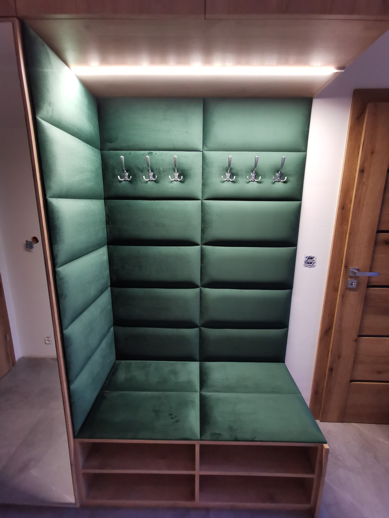 Wnęka z siedziskiem i wieszakami, obita zielonym, pikowanym welurem, oświetlona od góry, z drewnianą półką na buty i jasną podłogą.