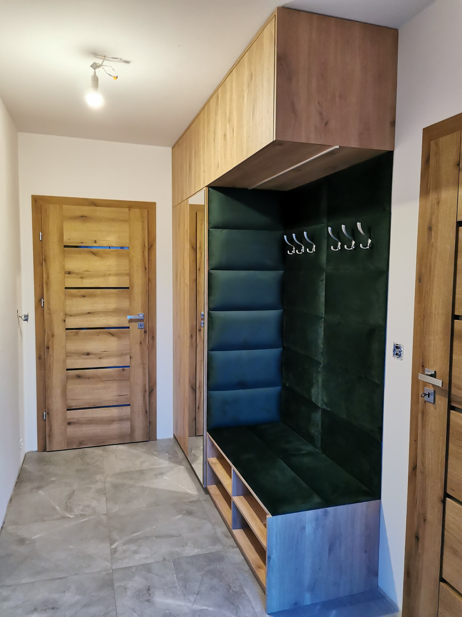 Przedpokój z zabudową meblową w odcieniach drewna i zieleni, z miękkim siedziskiem i wieszakami, oświetlony pojedynczą żarówką na suficie i wykończony płytkami imitującymi marmur.