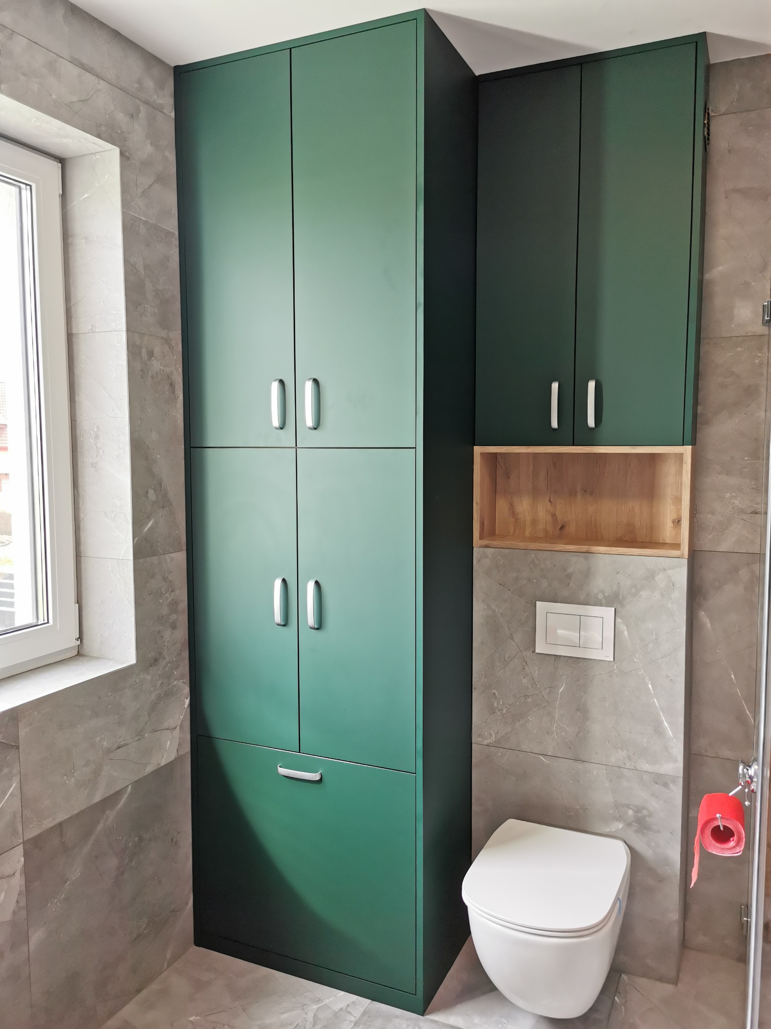Wysoka, zielona szafa łazienkowa z szufladą i srebrnymi uchwytami, zamontowana obok toalety i drewnianej półki w nowoczesnej łazience wykończonej płytkami imitującymi marmur.