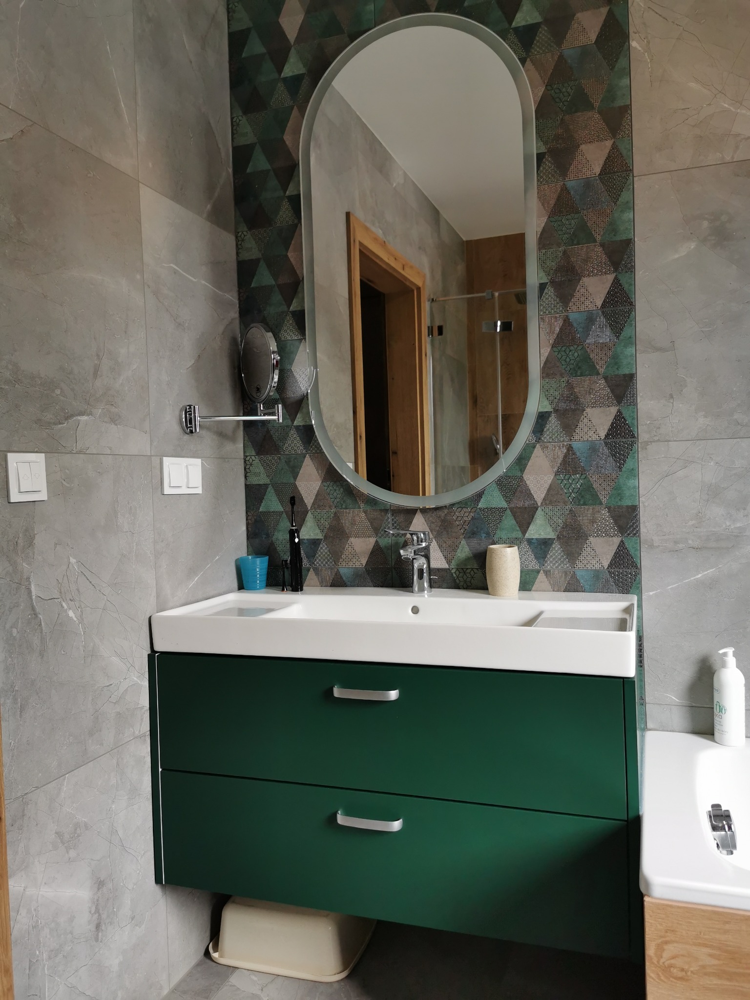 Nowoczesna łazienka z zieloną szafką pod umywalką, owalnym lustrem i geometryczną tapetą w odcieniach szarości, zieleni i brązu.