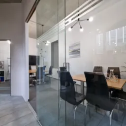 Nowoczesne biuro z szklanymi ścianami, widoczna sala konferencyjna z drewnianym stołem, czarnymi krzesłami, rośliną doniczkową i minimalistyczną lampą sufitową.