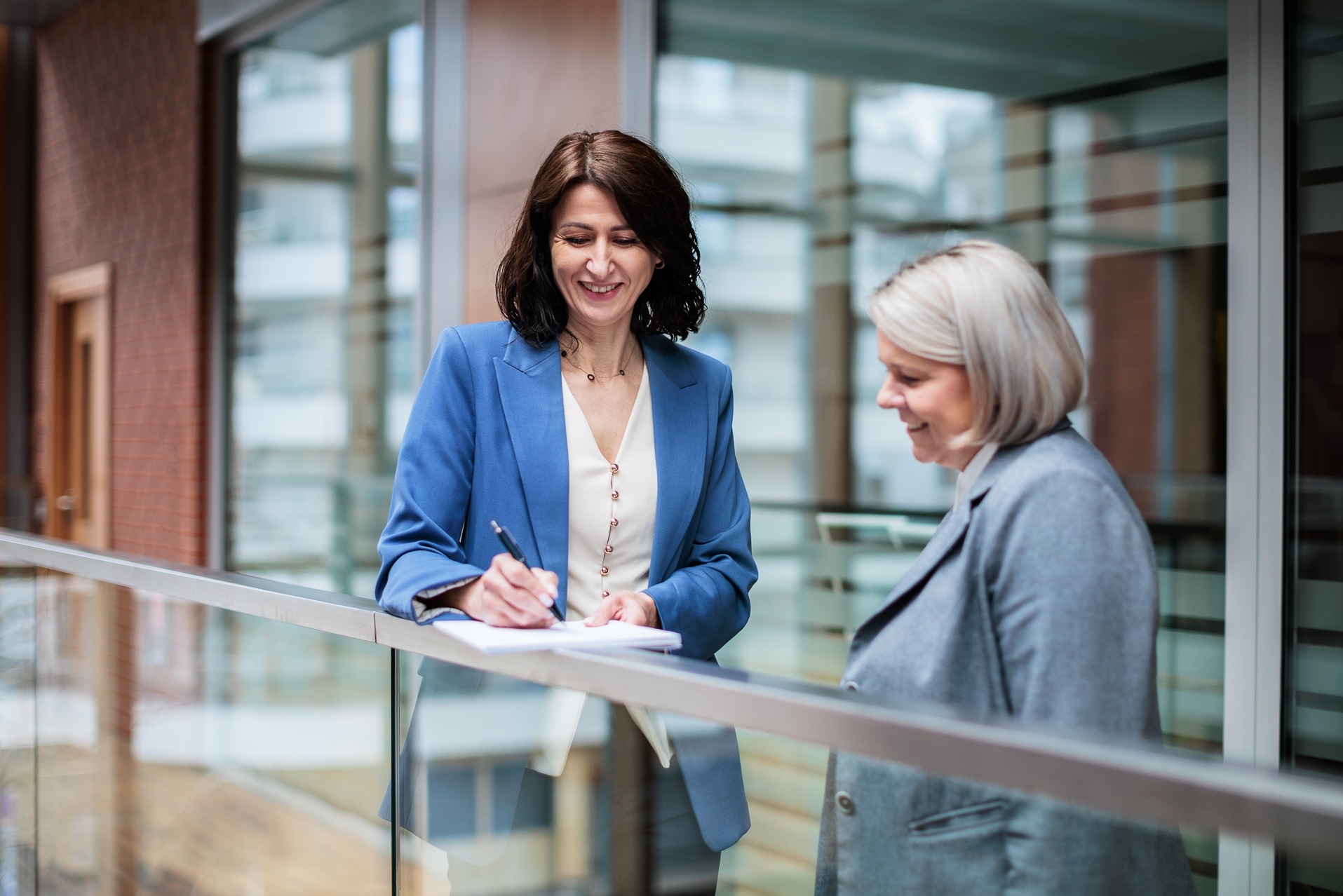 Kobieta w niebieskim żakiecie podpisuje dokument, uśmiechając się do drugiej kobiety w szarym żakiecie. Rozmowa w jasnym, nowoczesnym wnętrzu.